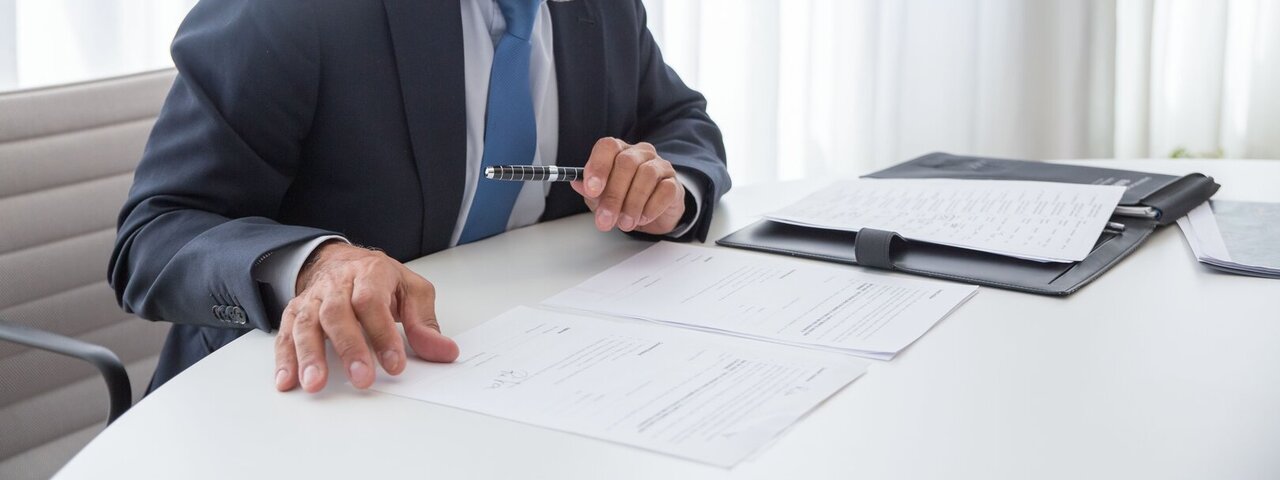 A professional in his mid-50s in a dark suit and glasses reviewing papers at an office desk