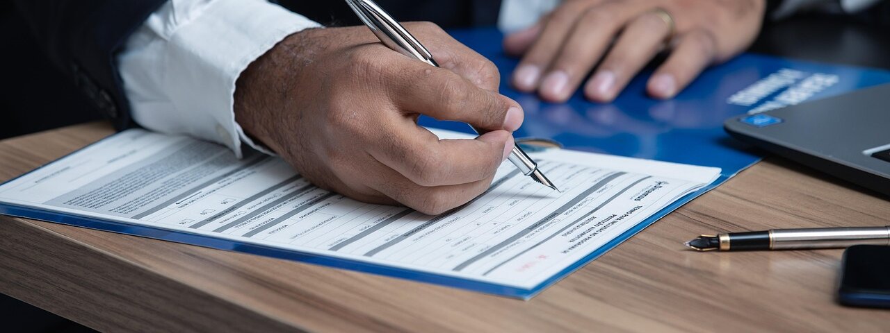 A professional in a business suit signing a printed form at a wooden desk, with a laptop nearby.