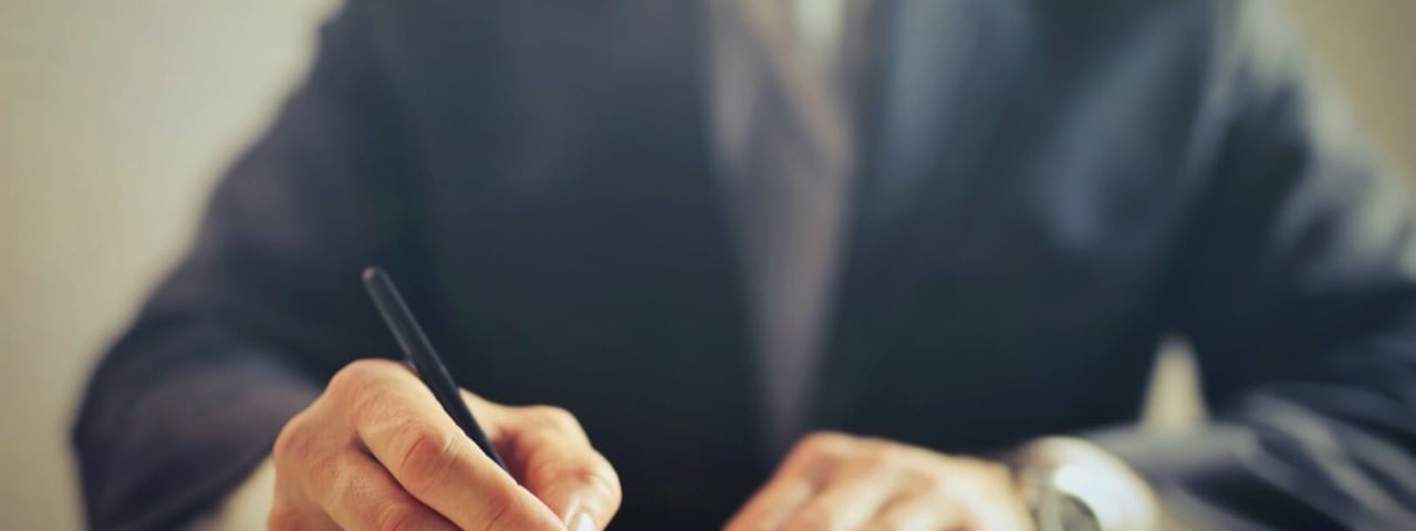 A professional in a dark suit holds a pen above a blank document on a white desk; face not shown.