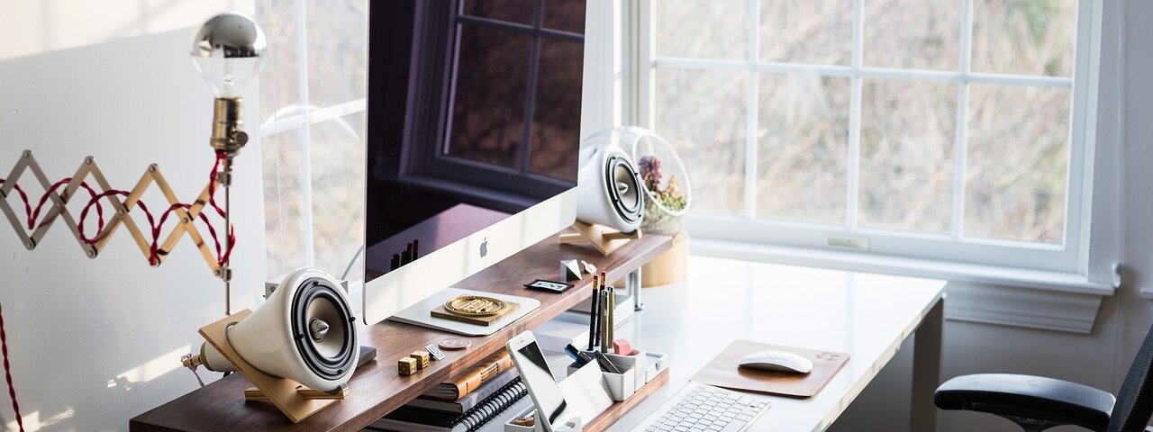 A tidy home office desk with a desktop computer, notebook, and desk lamp beside a window with natural light.