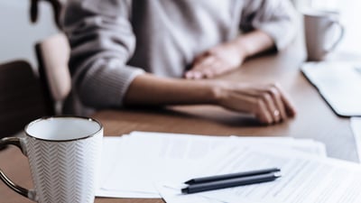 A person's hands resting on a wooden kitchen table beside a stack of paperwork, a pen, and a coffee mug, reviewing documents at home.