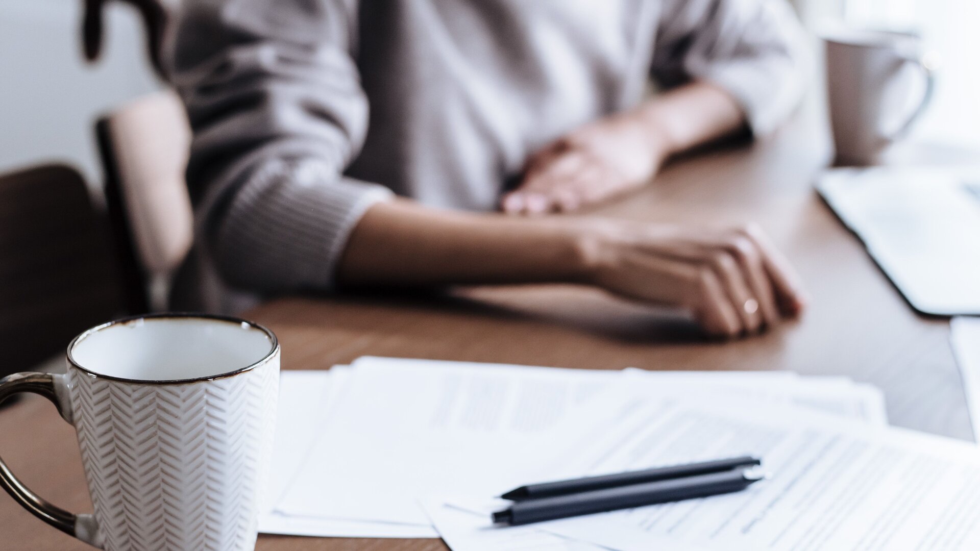 Household paperwork, a pen, and a coffee mug on a wooden kitchen table, with a person seated at the table in daylight.
