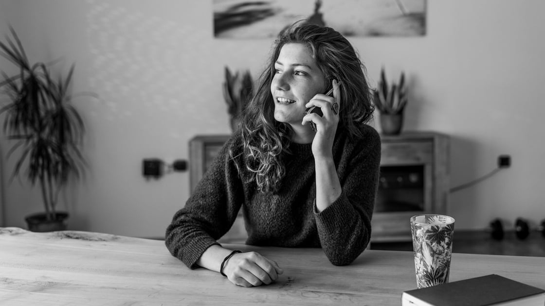 Mujer en la mesa de la cocina hablando por teléfono mientras revisa notas escritas a mano, fotografía en blanco y negro.