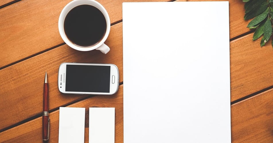 Overhead view of a warm wooden desk with a coffee mug, a smartphone, a pen, and a blank sheet of paper.