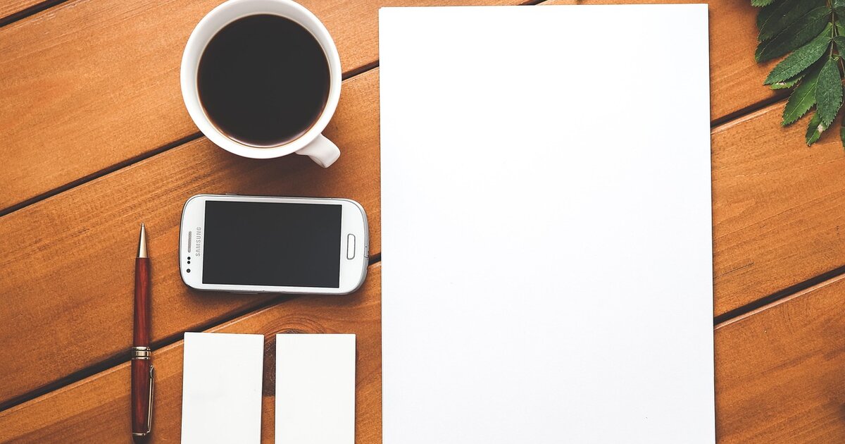 Overhead view of a warm wooden desk with a coffee mug, a smartphone, a pen, and a blank sheet of paper.