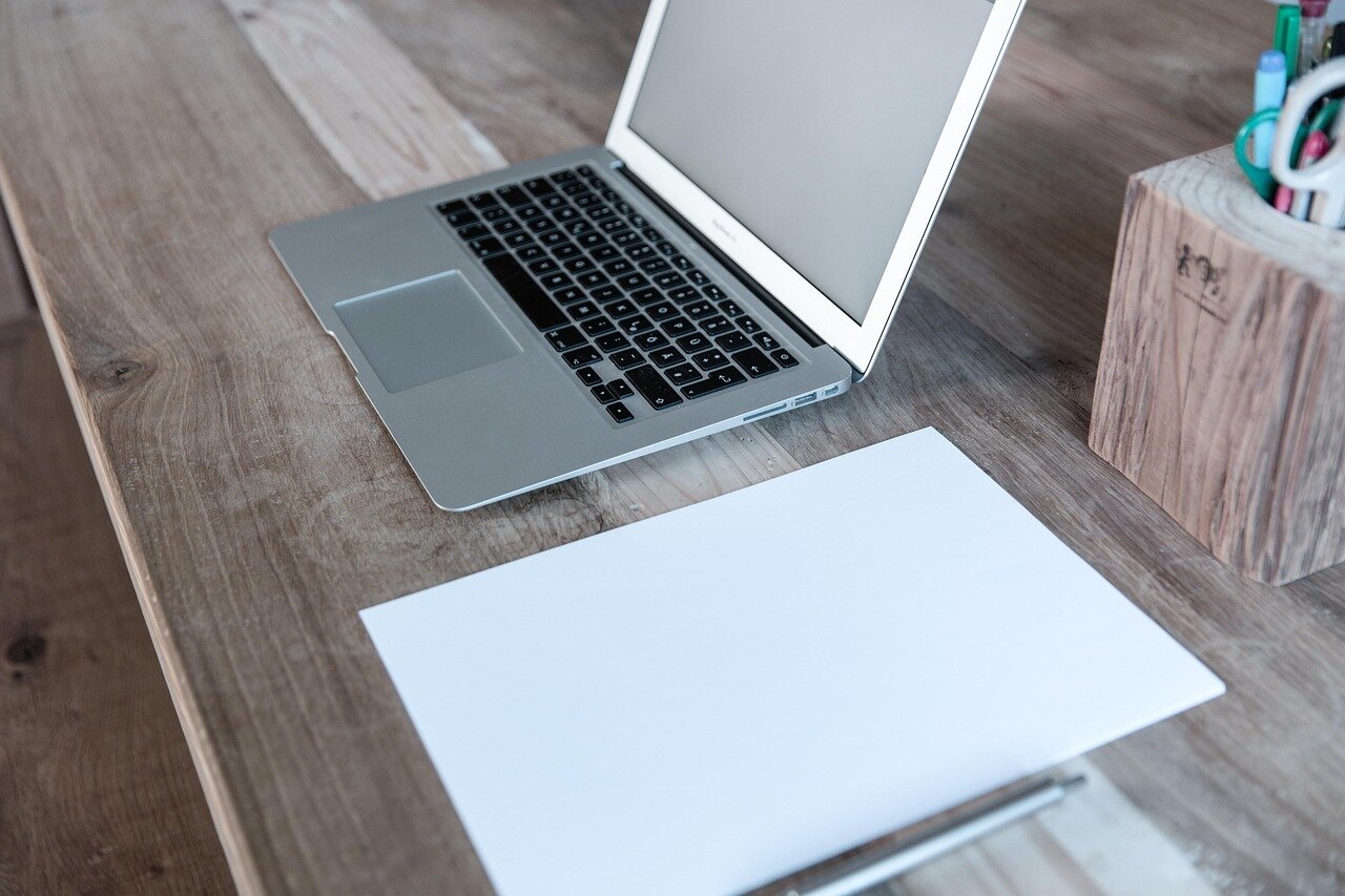 A laptop and a blank sheet of paper with a pen on a wooden desk.