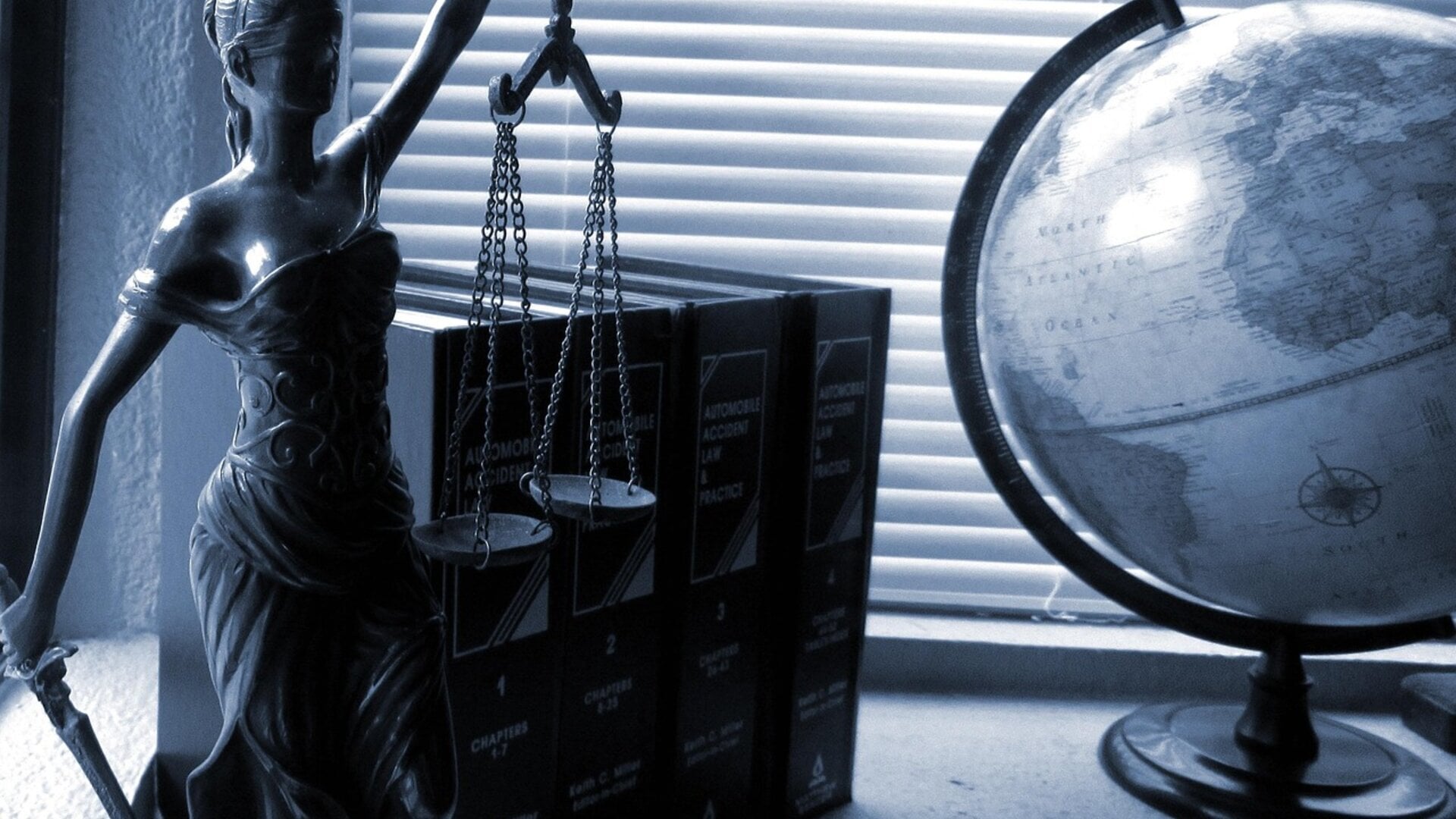 Scales of justice statue beside law books and a world globe, photographed in deep blue tones