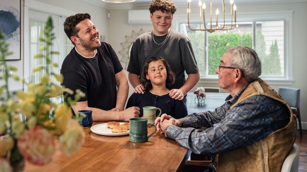Three generations of a family — a father, teenage son, young daughter, and grandfather — laughing together around a dining table at home.